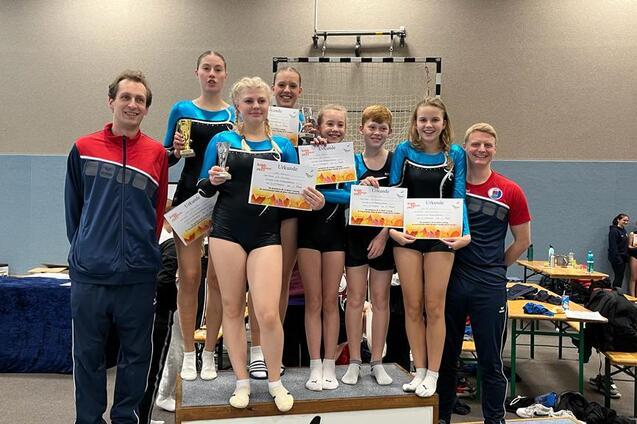 Gruppenszene mit Gymnastikern, die Pokale und Urkunden halten, umgeben von Trainern in einer Sporthalle.