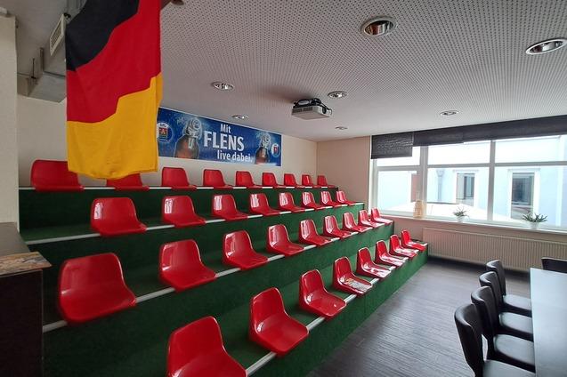 Rote Sitzpl&auml;tze auf einer gr&uuml;nen Trib&uuml;ne in einem Raum mit Fenster und deutscher Flagge an der Wand.