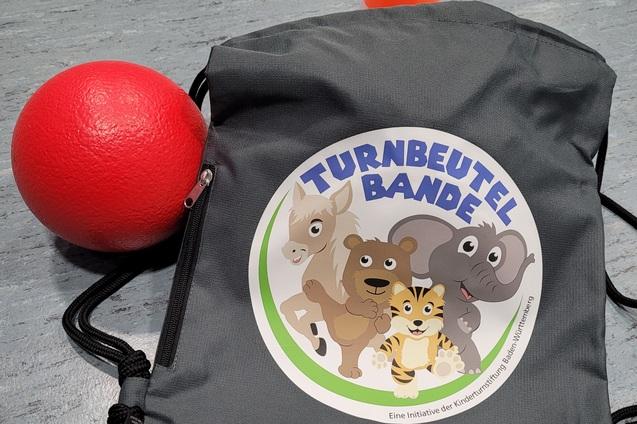 Grauer Turnbeutel mit buntem Aufdruck, rotem Ball und konischer Warnmarkierung auf einem grauen Untergrund.