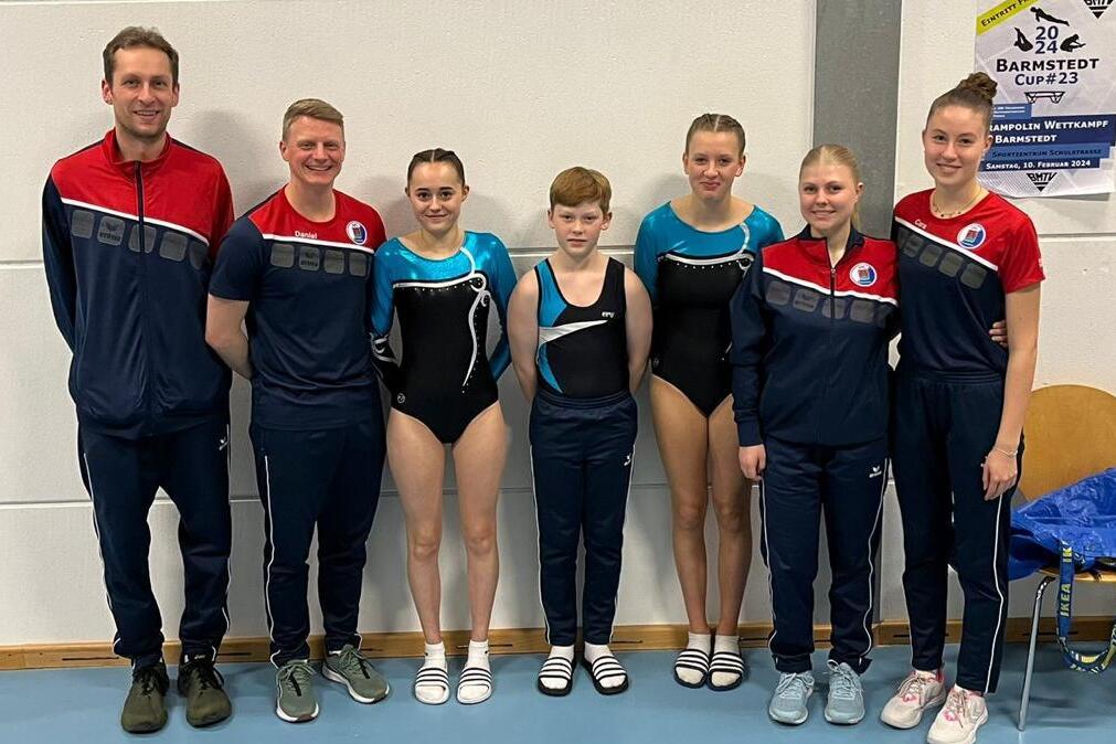 Gruppe von Gymnastikern und Trainern in Sportkleidung, l&auml;chelnd vor einer Wand mit Wettkampfinformationen.