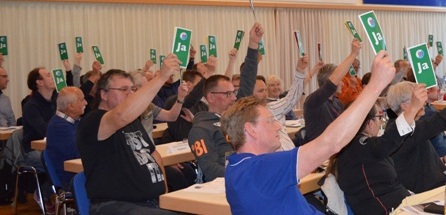 Versammlungsteilnehmer heben gr&uuml;ne &bdquo;Ja&ldquo;-Stimmkarten in die H&ouml;he, w&auml;hrend sie an langen Tischen sitzen.