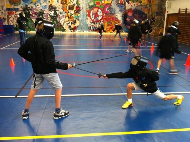 Zwei Kinder im Fencing-Anzug fencen auf einer Sporthalle mit farbenfrohem Graffiti-Hintergrund und orangefarbenen Hütchen.