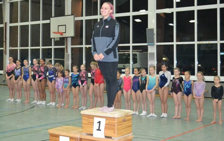 Trainerin steht auf einem Siegertreppchen, umgeben von gymnastischen Kindern in Sportanzügen in einer Halle.