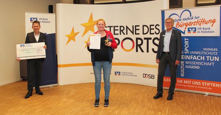 Siegerehrung Sterne des Sports Drei Personen stehen in einem Raum vor einem Banner, eine Person hält eine Urkunde und einen Pokal.