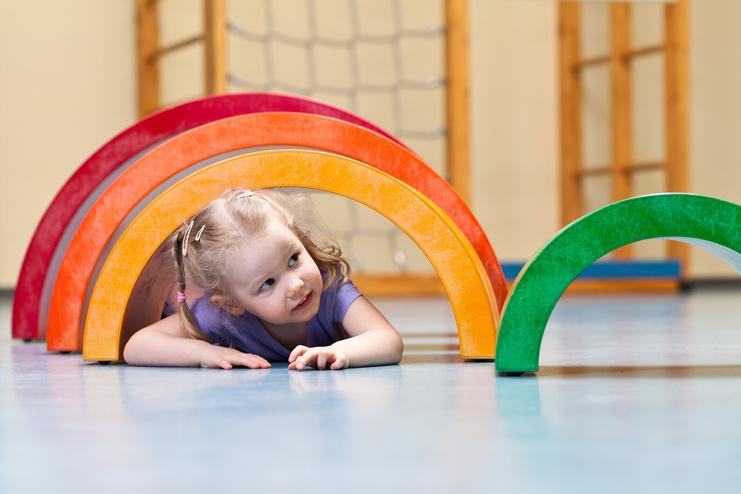 Mädchen krabbelt unter bunten Holzbögen in einer Spielhalle auf dem Boden.