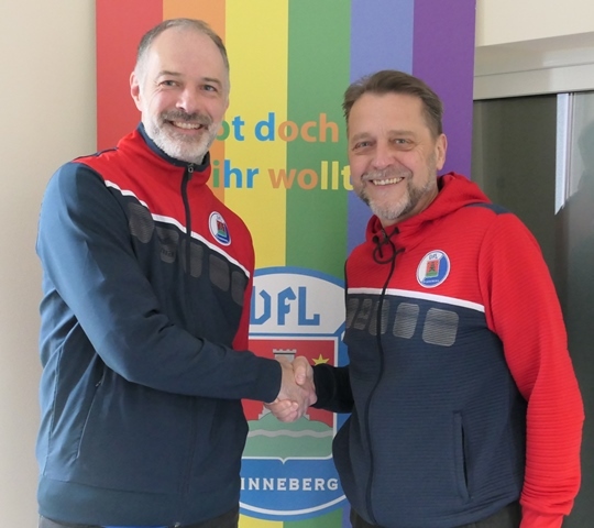 Zwei M&auml;nner sch&uuml;tteln sich die H&auml;nde vor einem bunten Banner mit der Aufschrift "UFL". Beide tragen sportliche Jacken.