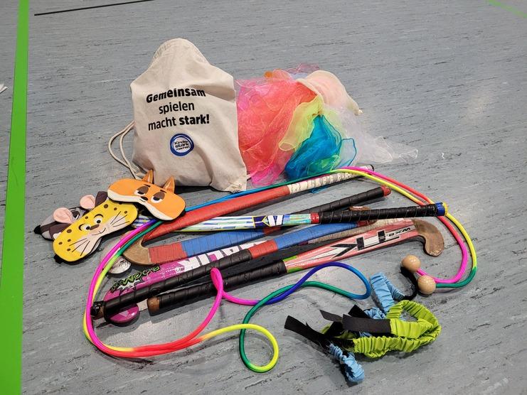 Beutel mit Spielmaterialien, darunter Masken, Stöcke und bunte Bänder auf grauem Boden. Text: „Gemeinsam spielen macht stark!“
