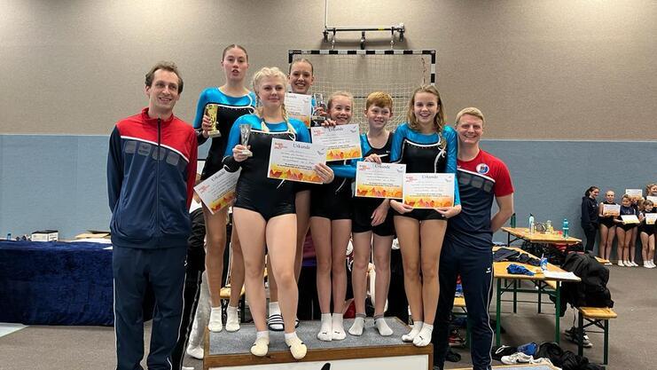 Gruppenszene mit Gymnastikern, die Pokale und Urkunden halten, umgeben von Trainern in einer Sporthalle.