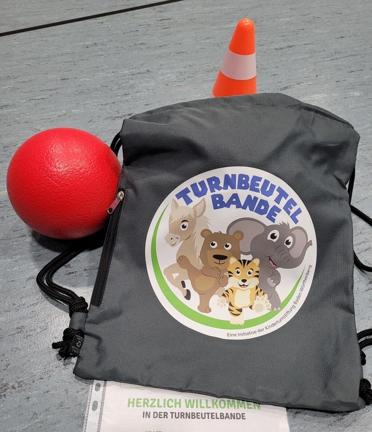 Grauer Turnbeutel mit buntem Aufdruck, rotem Ball und konischer Warnmarkierung auf einem grauen Untergrund.