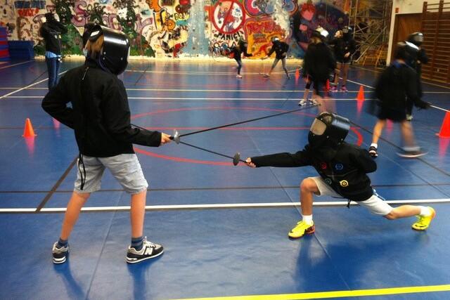 Zwei Kinder im Fencing-Anzug fencen auf einer Sporthalle mit farbenfrohem Graffiti-Hintergrund und orangefarbenen H&uuml;tchen.