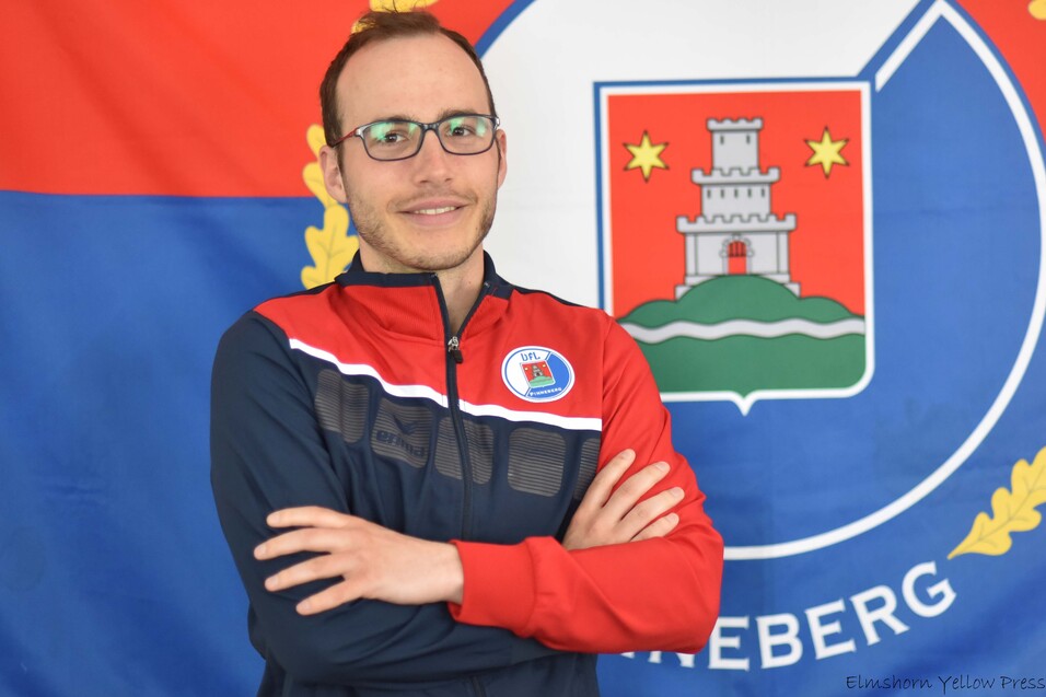 Mann mit Brille in Sportjacke vor einer Fahne, die ein Wappen mit einem Schloss und bunten Symbolen zeigt.