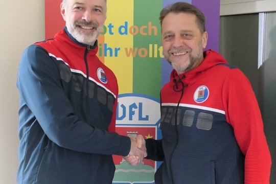 Zwei M&auml;nner sch&uuml;tteln sich die H&auml;nde vor einem bunten Banner mit der Aufschrift "UFL". Beide tragen sportliche Jacken.