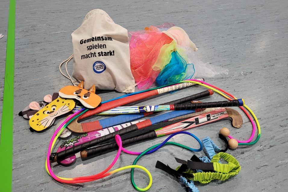 Beutel mit Spielmaterialien, darunter Masken, St&ouml;cke und bunte B&auml;nder auf grauem Boden. Text: &bdquo;Gemeinsam spielen macht stark!&ldquo;