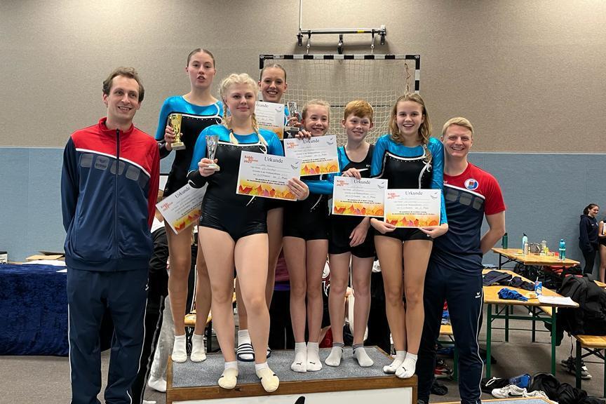 Gruppenszene mit Gymnastikern, die Pokale und Urkunden halten, umgeben von Trainern in einer Sporthalle.