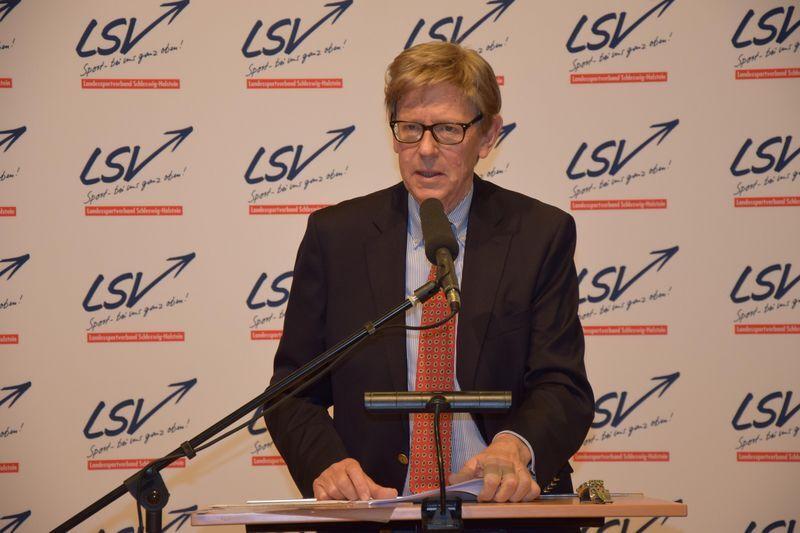 Mann mit Brille spricht am Mikrofon vor einem LSV-Logo auf einer Pr&auml;sentationswand.