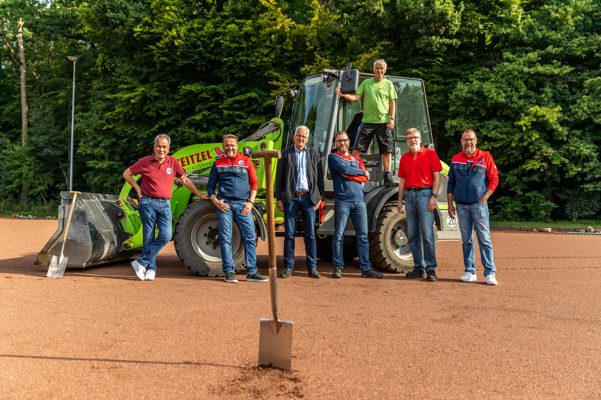 Gruppierung von sieben Personen, darunter ein Mann auf einem Bagger, alle auf einem roten Sportplatz.