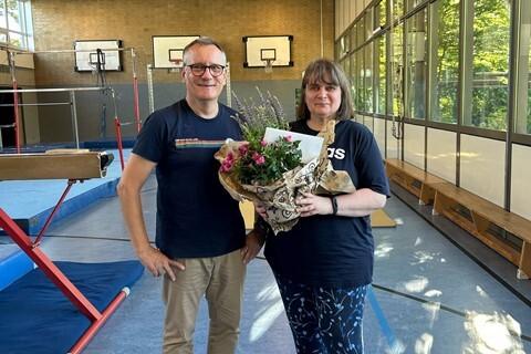 Zwei Personen stehen in einer Turnhalle und halten gemeinsam einen Blumenstrau&szlig;. Im Hintergrund sind Turnger&auml;te sichtbar.
