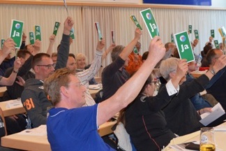 Versammlungsteilnehmer heben gr&uuml;ne &bdquo;Ja&ldquo;-Stimmkarten in die H&ouml;he, w&auml;hrend sie an langen Tischen sitzen.