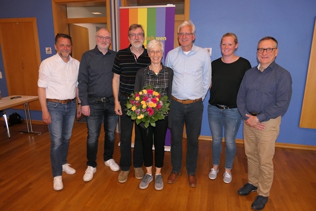 Gruppe von sechs Personen mit Blumenstrau&szlig;, stehend vor einer bunten Wand im Raum.