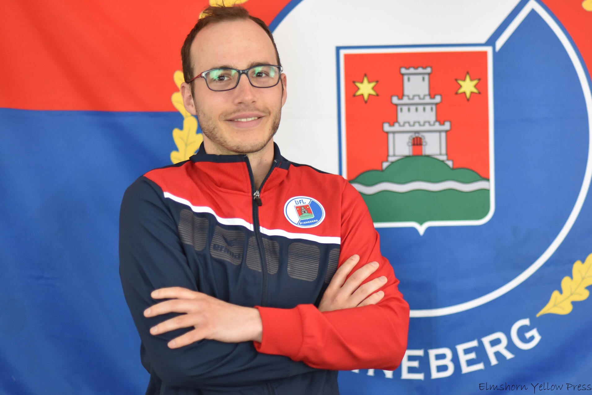 Mann mit Brille in Sportjacke vor einer Fahne, die ein Wappen mit einem Schloss und bunten Symbolen zeigt.