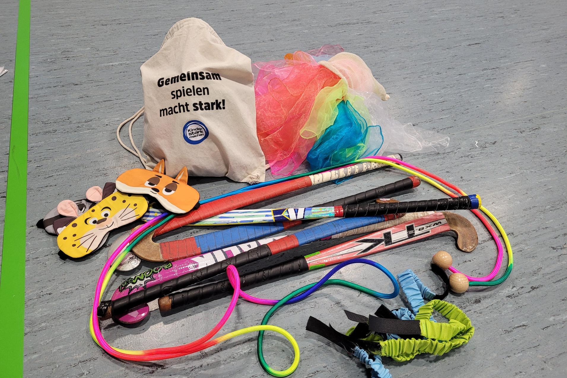 Beutel mit Spielmaterialien, darunter Masken, Stöcke und bunte Bänder auf grauem Boden. Text: „Gemeinsam spielen macht stark!“