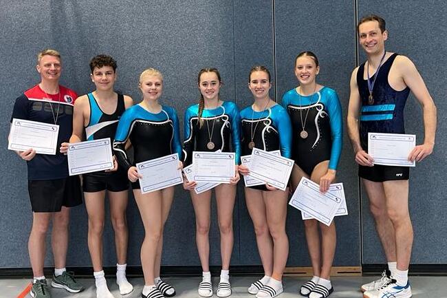 Gruppe von sieben Personen in Sportkleidung holding Urkunden und Medaillen, stehend vor einer Sportwand.