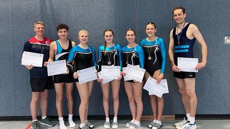 Gruppe von sieben Personen in Sportkleidung holding Urkunden und Medaillen, stehend vor einer Sportwand.