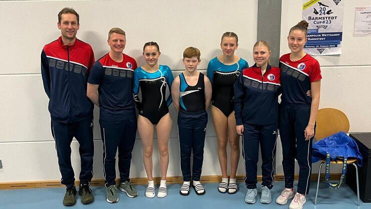 Gruppe von Gymnastikern und Trainern in Sportkleidung, lächelnd vor einer Wand mit Wettkampfinformationen.