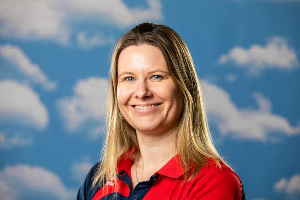 Portrait einer lächelnden Frau mit langen blonden Haaren, im roten und blauen Polohemd vor einem wolkigen blauen Hintergrund.