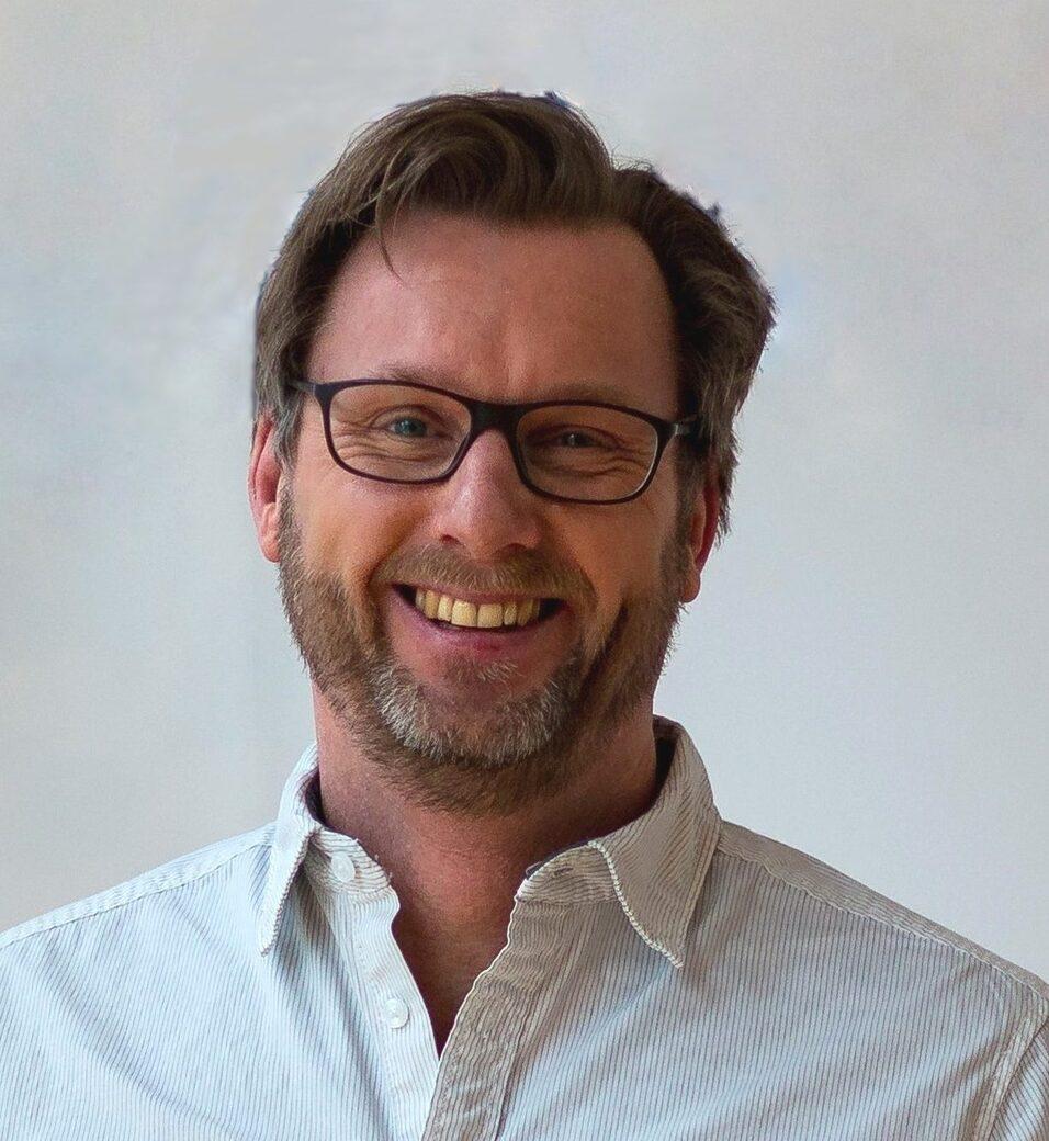 Nahaufnahme eines lächelnden Mannes mit Brille, kurzem Bart und einem hellen Hemd vor einer neutralen Hintergrundwand.