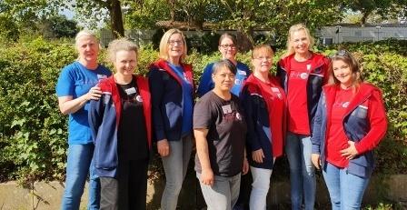 Gruppe von acht Frauen in roten und blauen T-Shirts, posierend vor einer grünen Hecke im Freien.