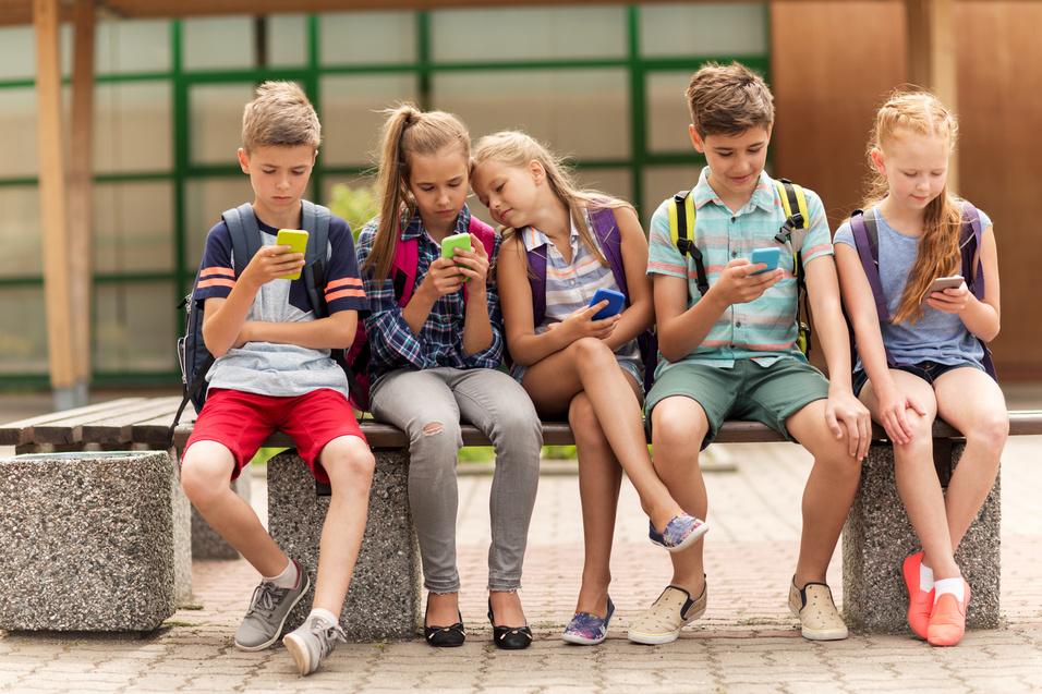 Fünf Kinder sitzen auf einer Bank und schauen auf ihre Smartphones, während sie verschiedene Apps nutzen.