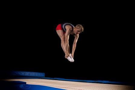 Athlet springt mit gestreckten Beinen auf einem Trampolin, schwarzer Hintergrund, in vorheriger Position.