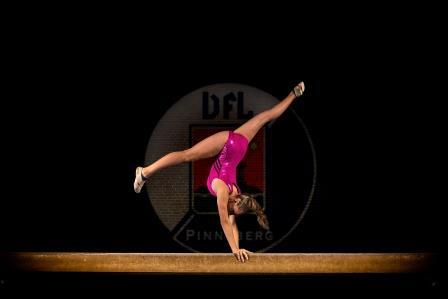 Turnerin in rosa Outfit, die auf einem Schwebebalken eine Handstand-Position einnimmt, vor schwarzem Hintergrund.