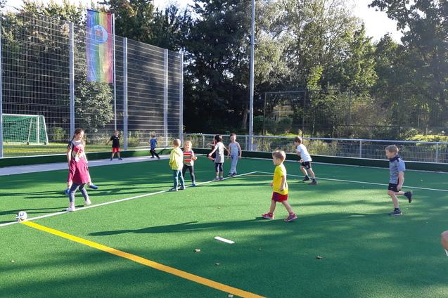 Gruppe von Kindern spielt auf einem Kunstrasenplatz, im Hintergrund sichtbar ein Fu&szlig;balltor und eine Regenbogenflagge.