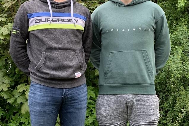 Zwei M&auml;nner stehen nebeneinander in einer gr&uuml;nen Umgebung, beide tragen sportliche Sweatshirts und Jeans.