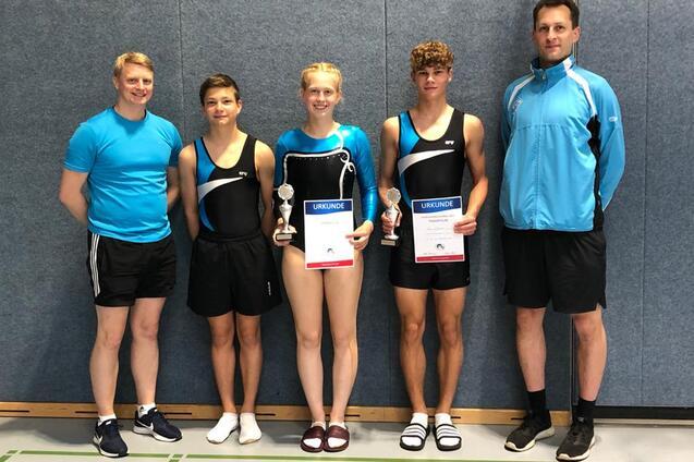 Gruppe von vier Sportlern in Kleidern f&uuml;r Turnen, halten Urkunden und Pokale, stehen vor einer grauen Wand.