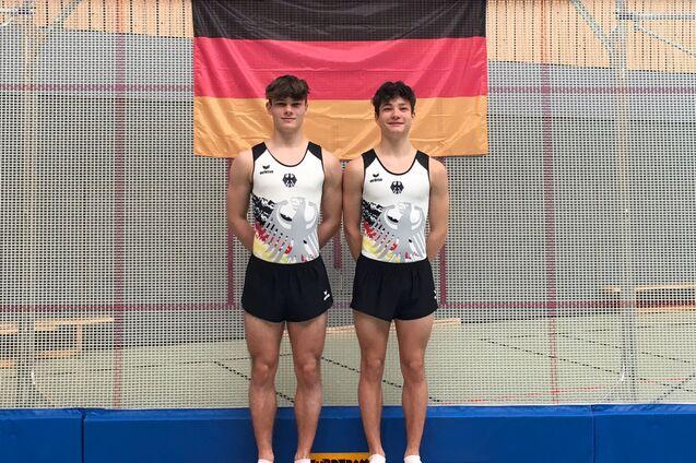 Zwei Turner in Trikots mit deutschem Adler posieren auf einer Trainingsmatte vor einer Deutschlandflagge.