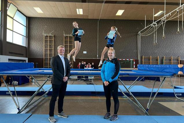 Drei Turnerinnen stehen auf einem Trampolin, w&auml;hrend eine von ihnen in der Luft schwebt und ein Dokument h&auml;lt.