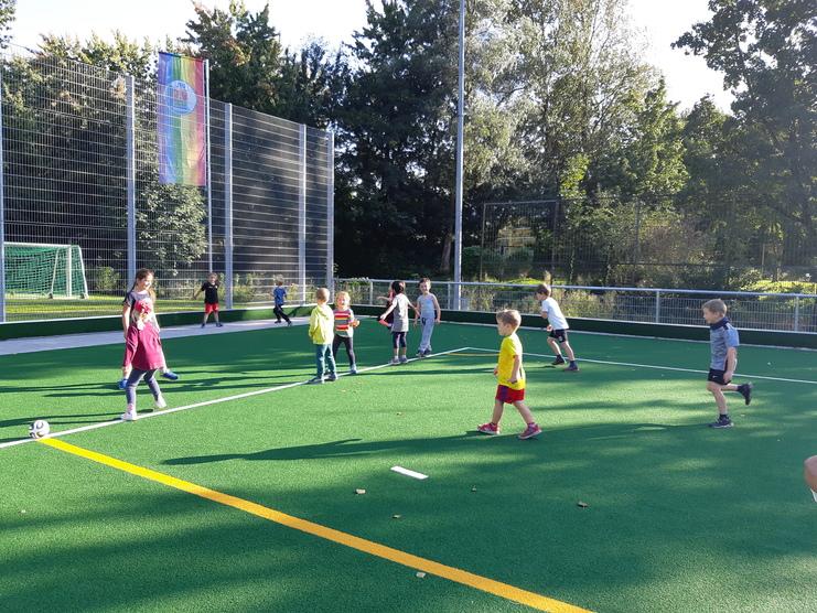 Gruppe von Kindern spielt auf einem Kunstrasenplatz, im Hintergrund sichtbar ein Fußballtor und eine Regenbogenflagge.