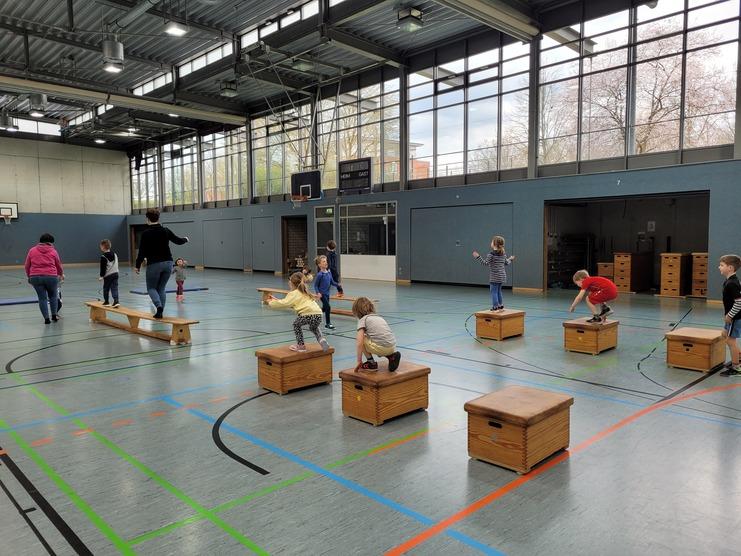 Kinder und Erwachsene üben Balancieren und Springen auf Holzkästen in einer Sporthalle mit großen Fenstern.