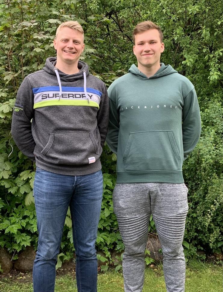 Zwei Männer stehen nebeneinander in einer grünen Umgebung, beide tragen sportliche Sweatshirts und Jeans.