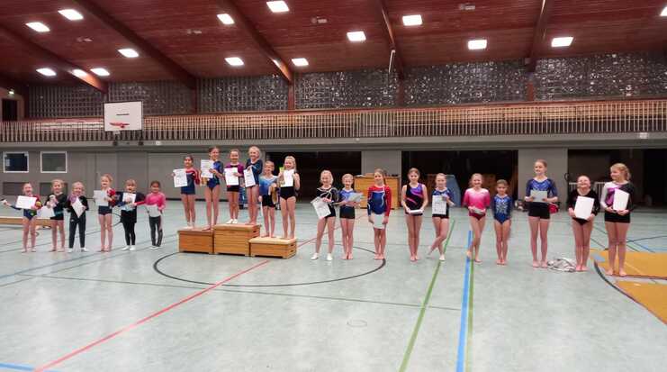 Gruppe junger Turnerinnen hält Urkunden in einer Turnhalle, einige stehen auf einem Podest.