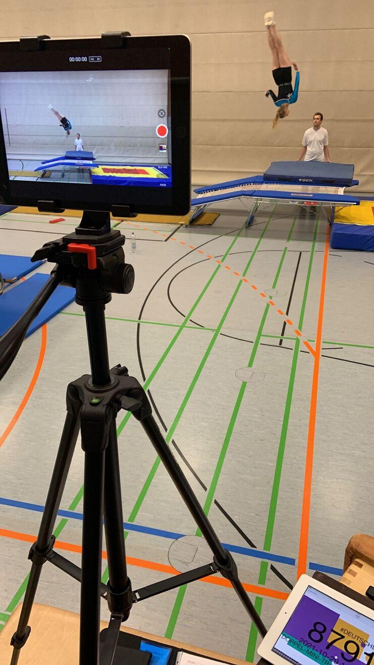 Auf einem Tischtennistraining sind ein Tisch, eine Kamera auf einem Stativ und ein Springer in der Luft zu sehen.
