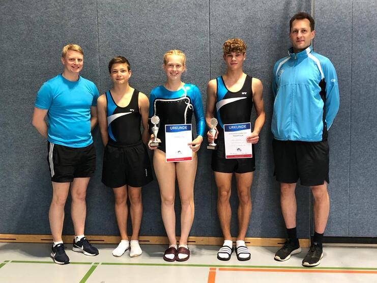 Gruppe von vier Sportlern in Kleidern für Turnen, halten Urkunden und Pokale, stehen vor einer grauen Wand.