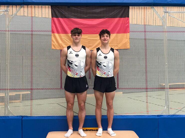 Zwei Turner in Trikots mit deutschem Adler posieren auf einer Trainingsmatte vor einer Deutschlandflagge.