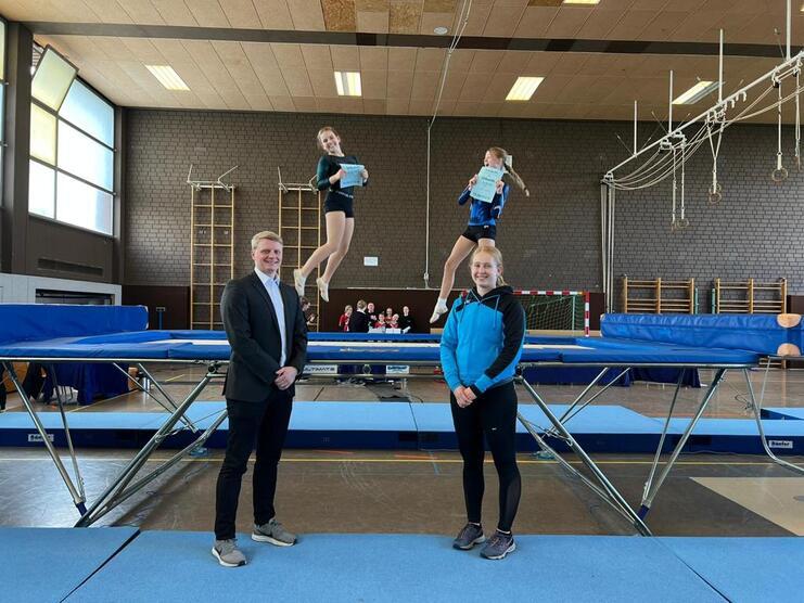 Drei Turnerinnen stehen auf einem Trampolin, während eine von ihnen in der Luft schwebt und ein Dokument hält.