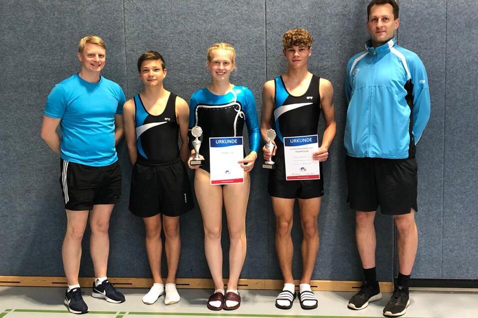 Gruppe von vier Sportlern in Kleidern f&uuml;r Turnen, halten Urkunden und Pokale, stehen vor einer grauen Wand.