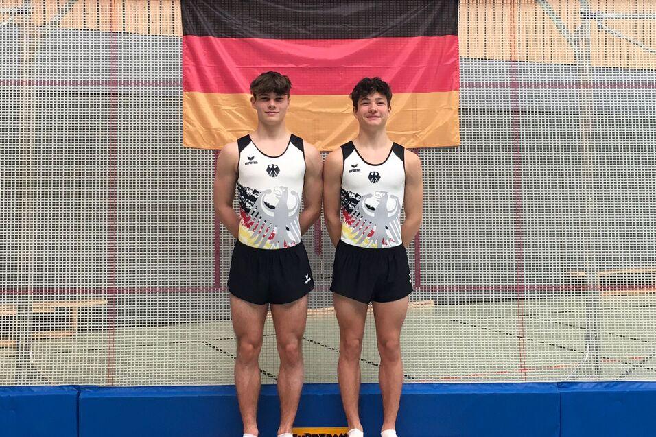 Zwei Turner in Trikots mit deutschem Adler posieren auf einer Trainingsmatte vor einer Deutschlandflagge.