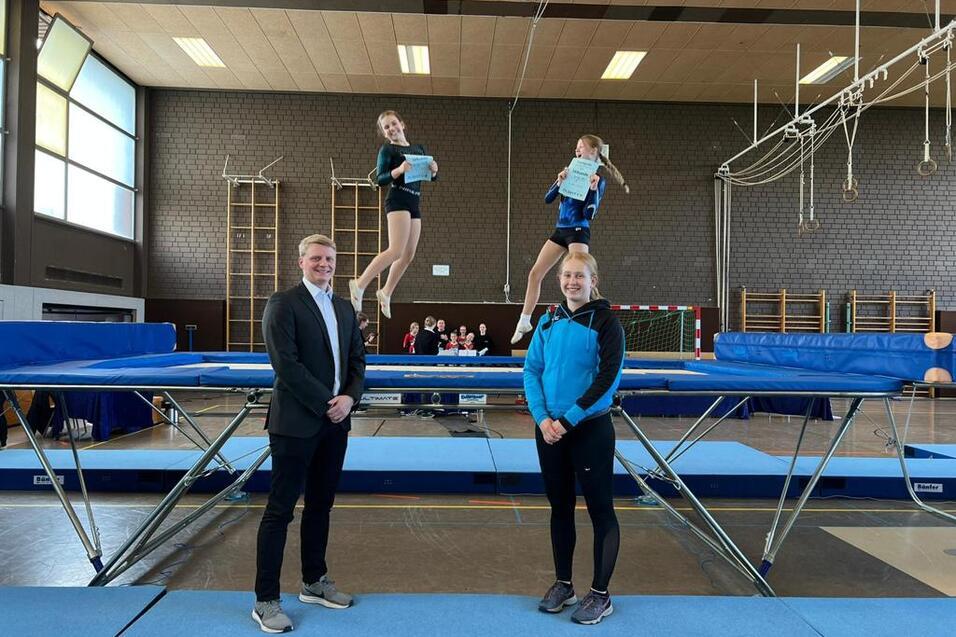 Drei Turnerinnen stehen auf einem Trampolin, w&auml;hrend eine von ihnen in der Luft schwebt und ein Dokument h&auml;lt.